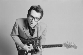 Becoming Elvis Costello: The Legend Goes In-Depth on His New ‘My Aim Is True’ Boxed Set, Origins of the Iconic Name and Glasses, and Finding a Sound and Persona That Transformed Rock Forever
